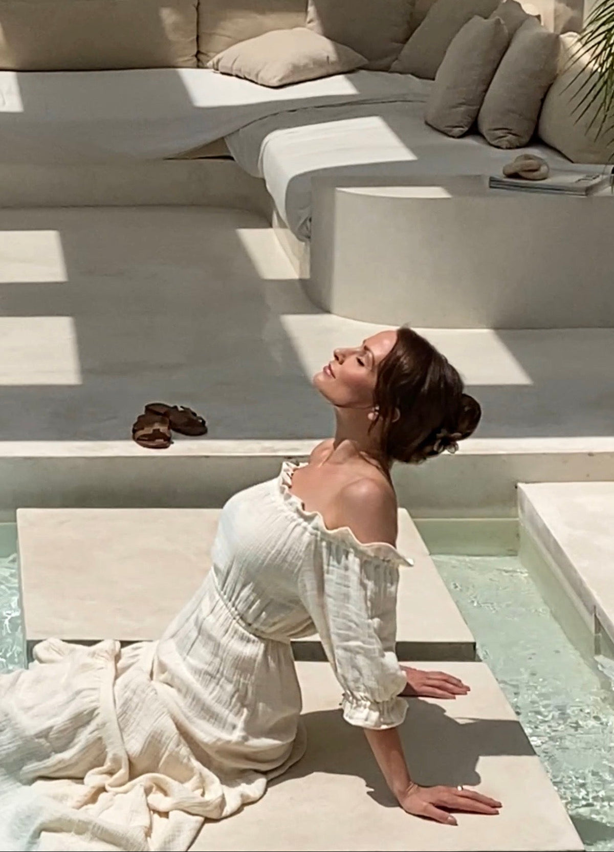Side profile of a woman with closed eyes bathing in the sun by a beige villa pool, wearing the natural off-shoulder dress made from organic cotton and hemp