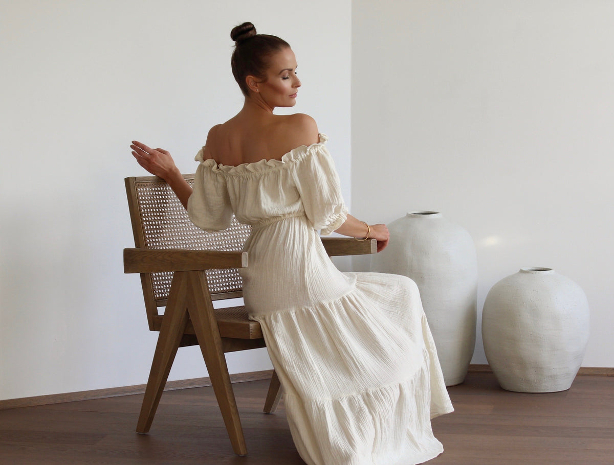 A woman seated on a minimalist wooden chair, captured from behind as she looks back over her shoulder with an elegant, sensual gaze while wearing the natural off-shoulder dress