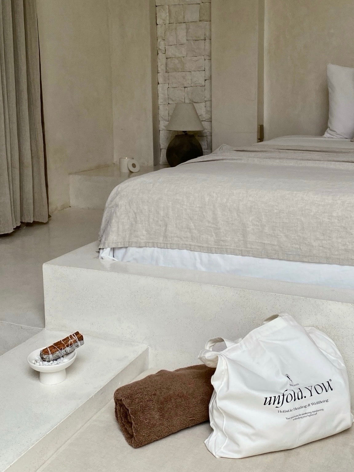 A white sage and cinnamon incense bundle resting in a heatproof dish on a bedside table in a serene bedroom, elegantly arranged alongside a soft linen tote bag and a textured cotton towel.