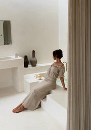 On-model upper-body view of the undyed luxury off-shoulder blouse showcasing its elastic ruffled neckline, shot on location sitting on a minimalist bathtub with light background.
