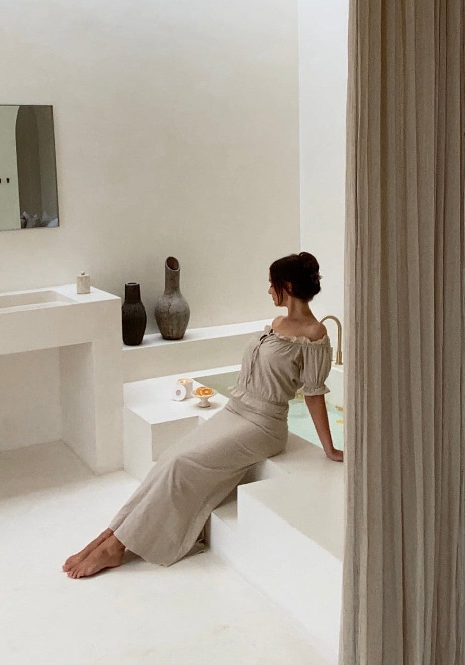 On-model upper-body view of the undyed luxury off-shoulder blouse showcasing its elastic ruffled neckline, shot on location sitting on a minimalist bathtub with light background.