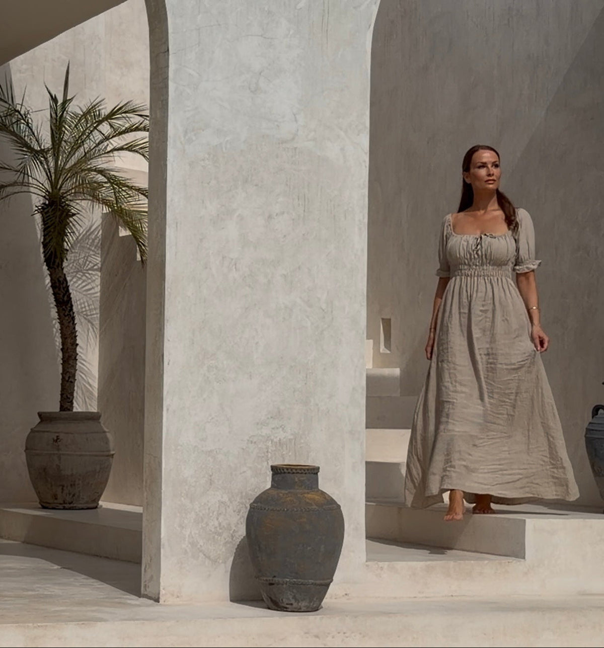 On-model view of a fluid, floor-length luxury resort dress in motion, captured inside a minimalist beige Bali villa with architectural palm tree details.