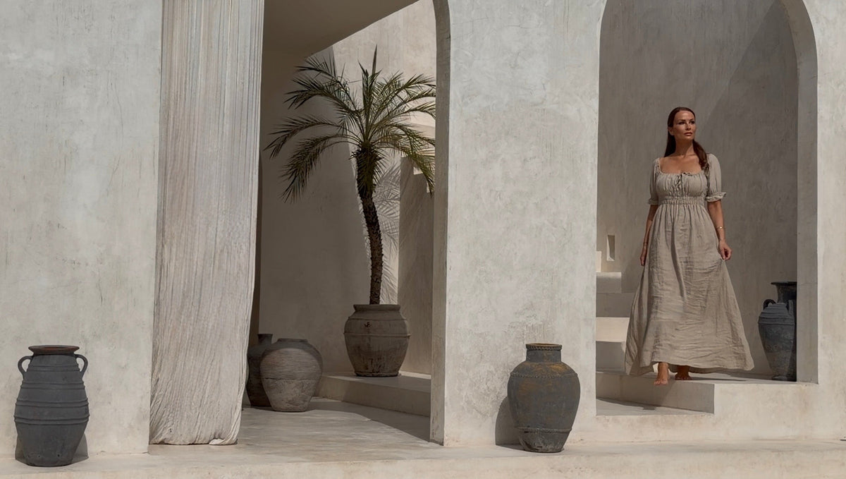 On-model view of a fluid, floor-length luxury resort dress in motion, captured inside a minimalist beige Bali villa with architectural palm tree details. 
