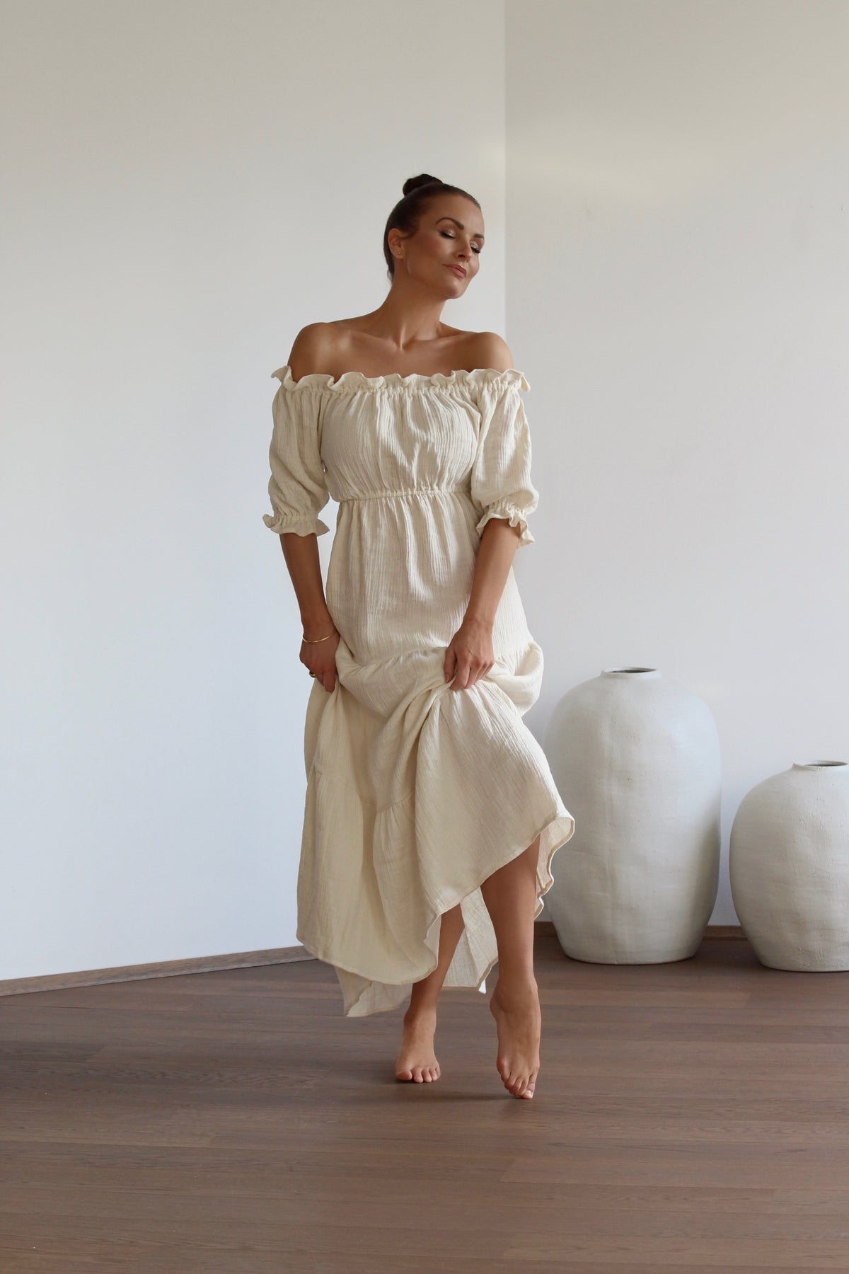 Full-length view of a woman standing in a serene, sunlit room, embodying a sensual presence while wearing the long natural undyed off-shoulder dress